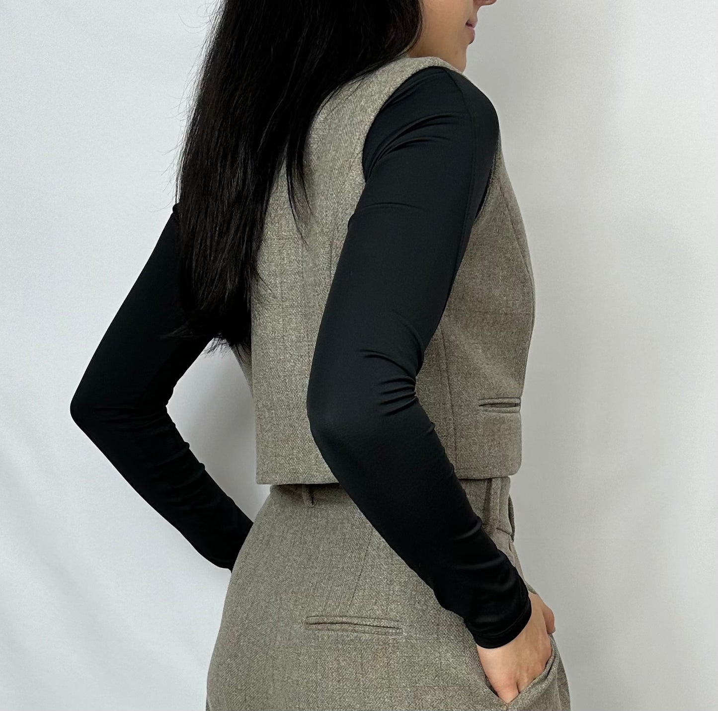 Model showcasing the back and side profile of a Sartorial Flair grey waistcoat paired with matching high-waisted trousers. The outfit is complemented by a black long-sleeve top, emphasizing the sleek design and tailored fit. This ensemble highlights the versatility of the suit for both formal and smart casual wear.