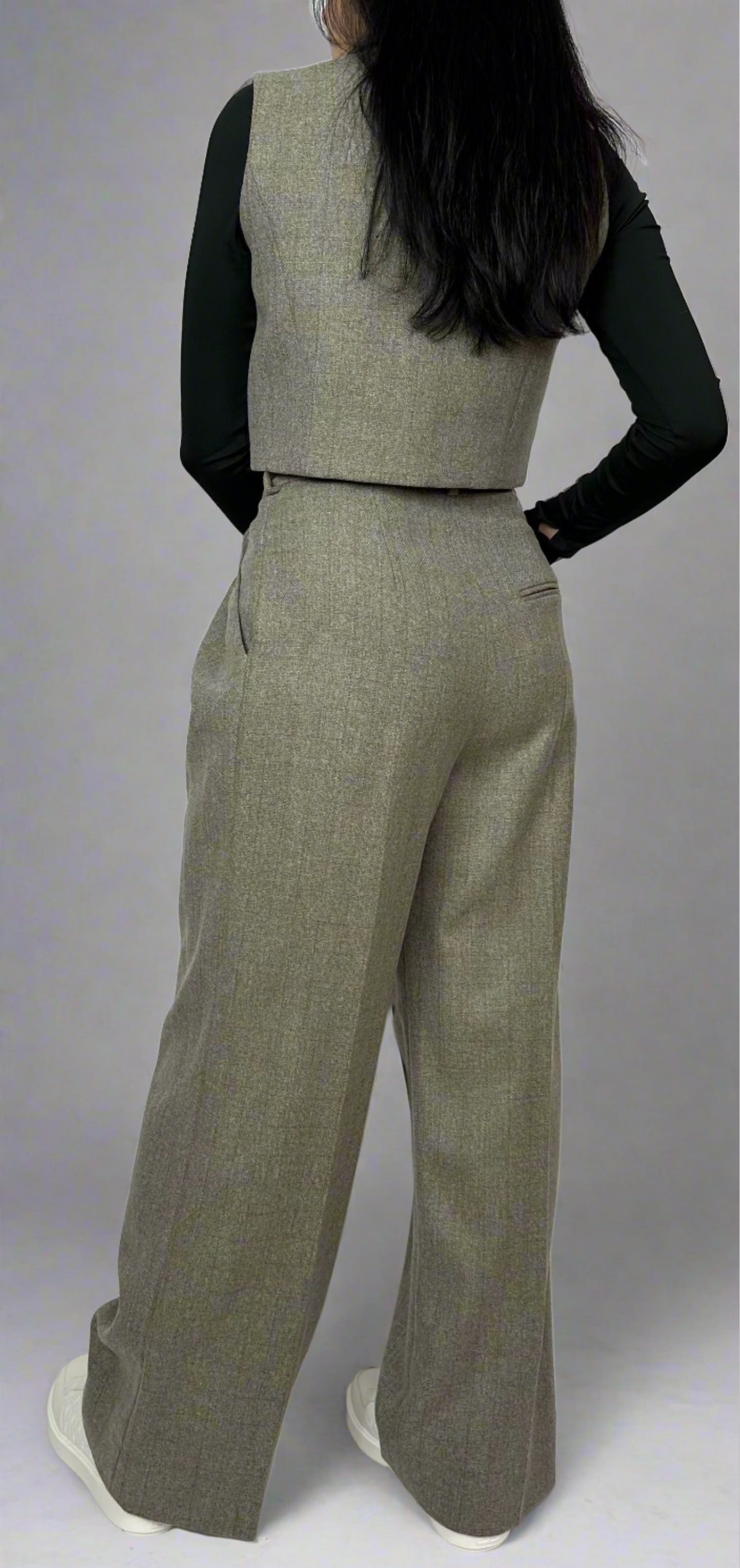 Model displaying the back profile of a Sartorial Flair grey waistcoat paired with matching high-waisted trousers. The outfit is complemented by a black long-sleeve top, emphasizing the tailored fit and sophisticated design. This ensemble highlights the versatility of the suit for both formal and smart casual wear.”