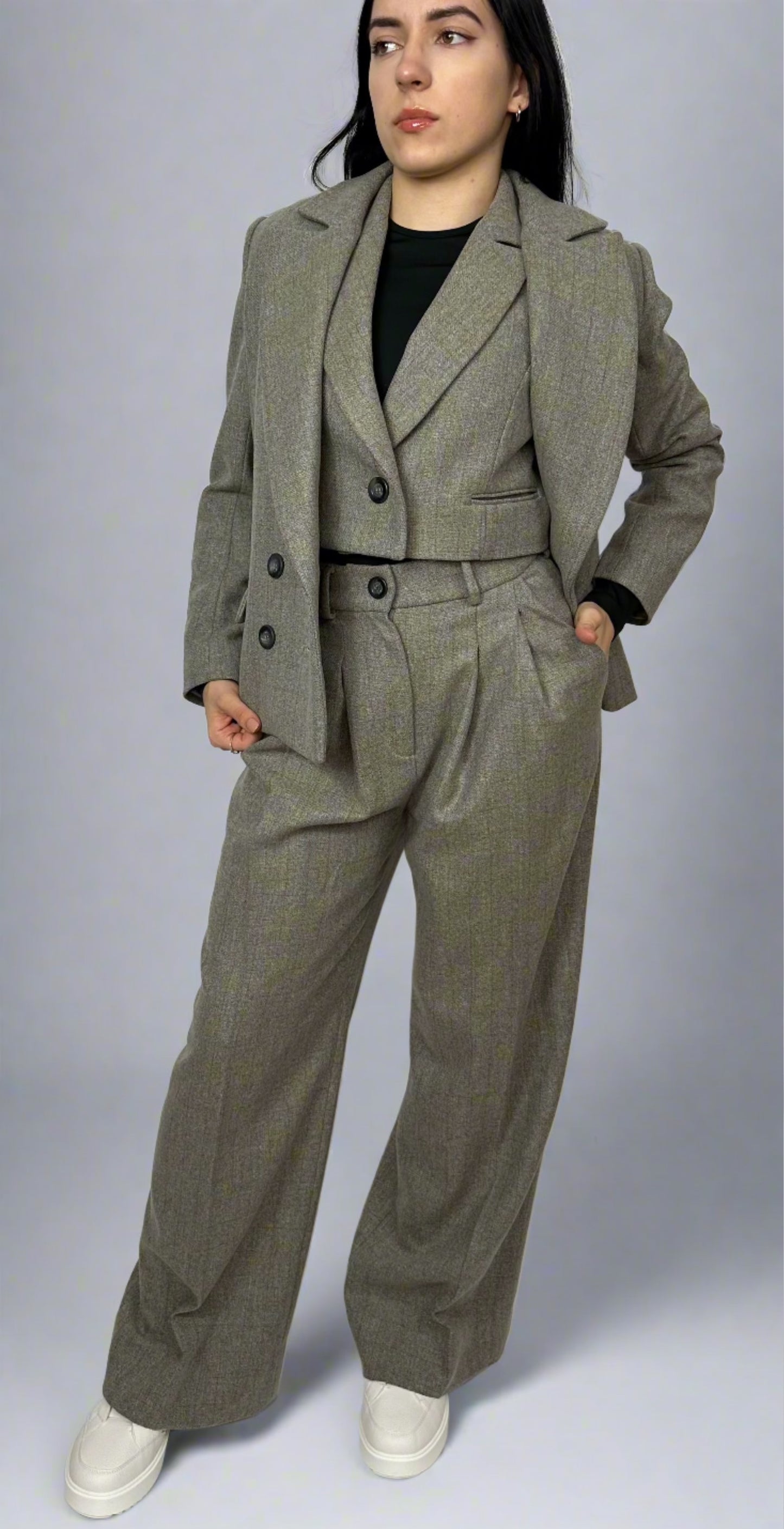 Model wearing Sartorial Flair grey three-piece suit, consisting of a double-breasted blazer, waistcoat with a single button, and high-waisted trousers. The outfit is paired with a black top and white trainers, showcasing how suits can be styled for both formal and smart casual looks.