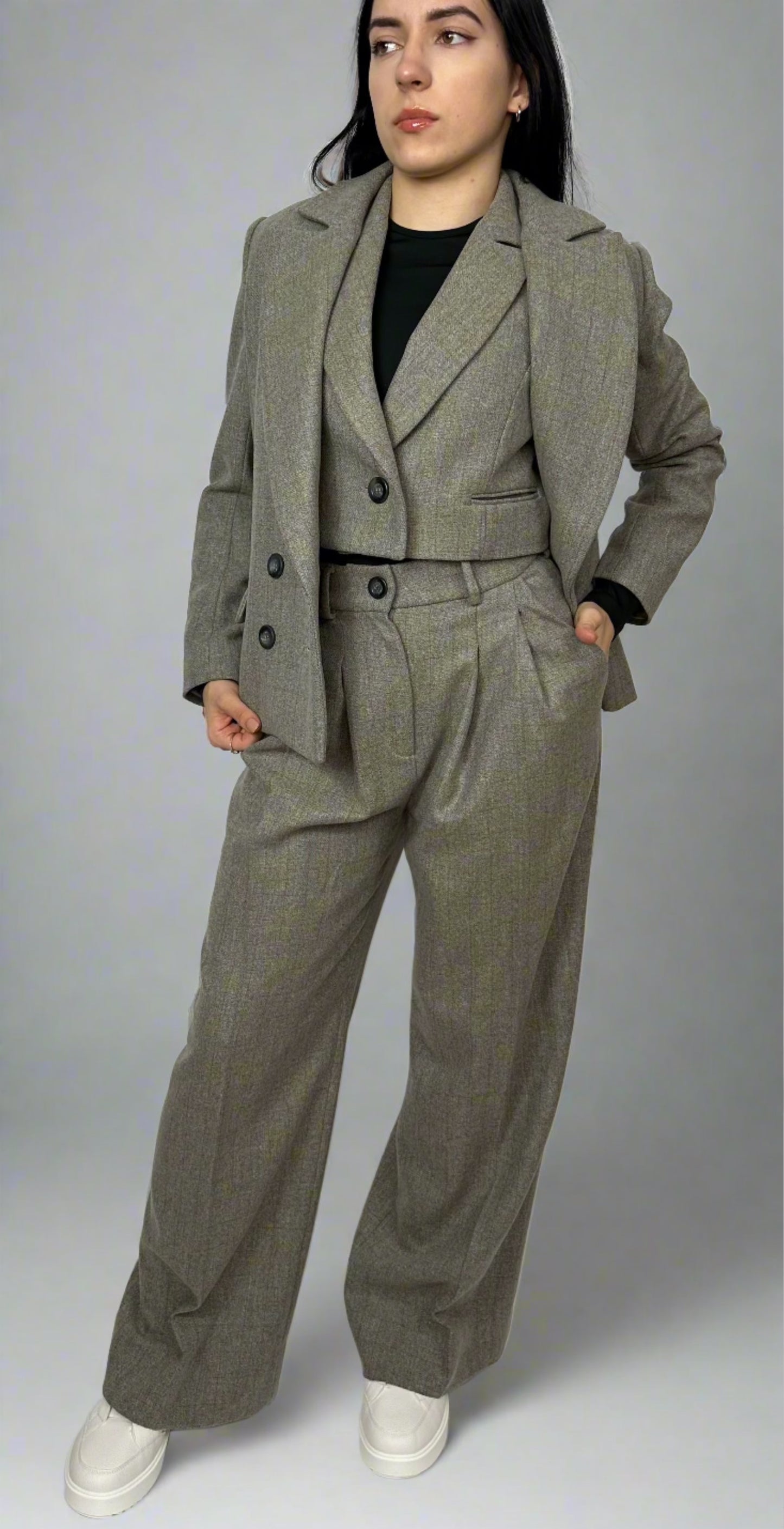 Model wearing Sartorial Flair grey three-piece suit, consisting of a double-breasted blazer, waistcoat with a single button, and high-waisted trousers. The outfit is paired with a black top and white trainers, showcasing how suits can be styled for both formal and smart casual looks.