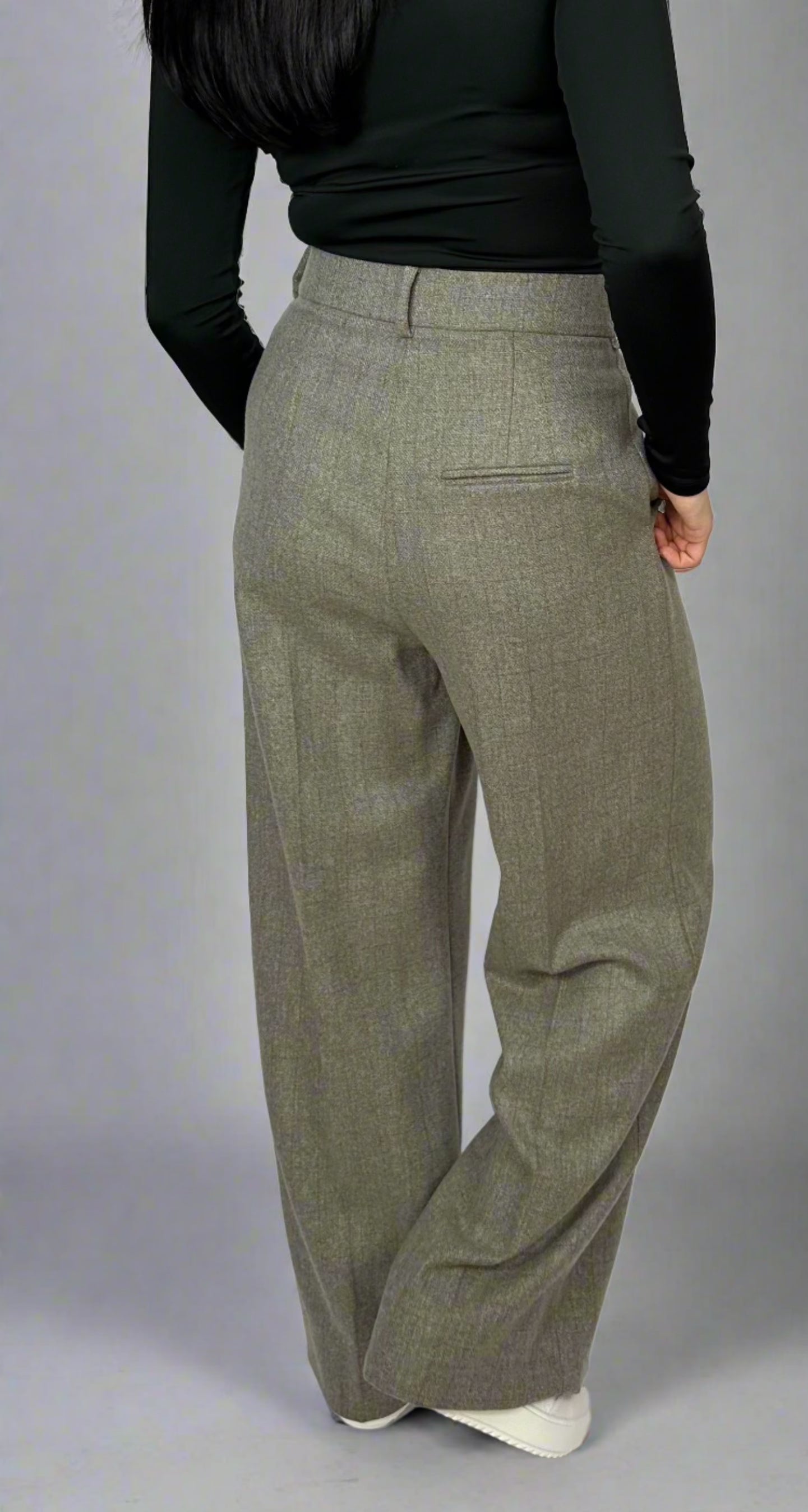 Rear view of a model wearing Sartorial Flair grey high-waisted trousers, paired with a black top. The back pocket detail and tailored fit showcase the trousers’ design, suitable for both formal and smart casual looks.