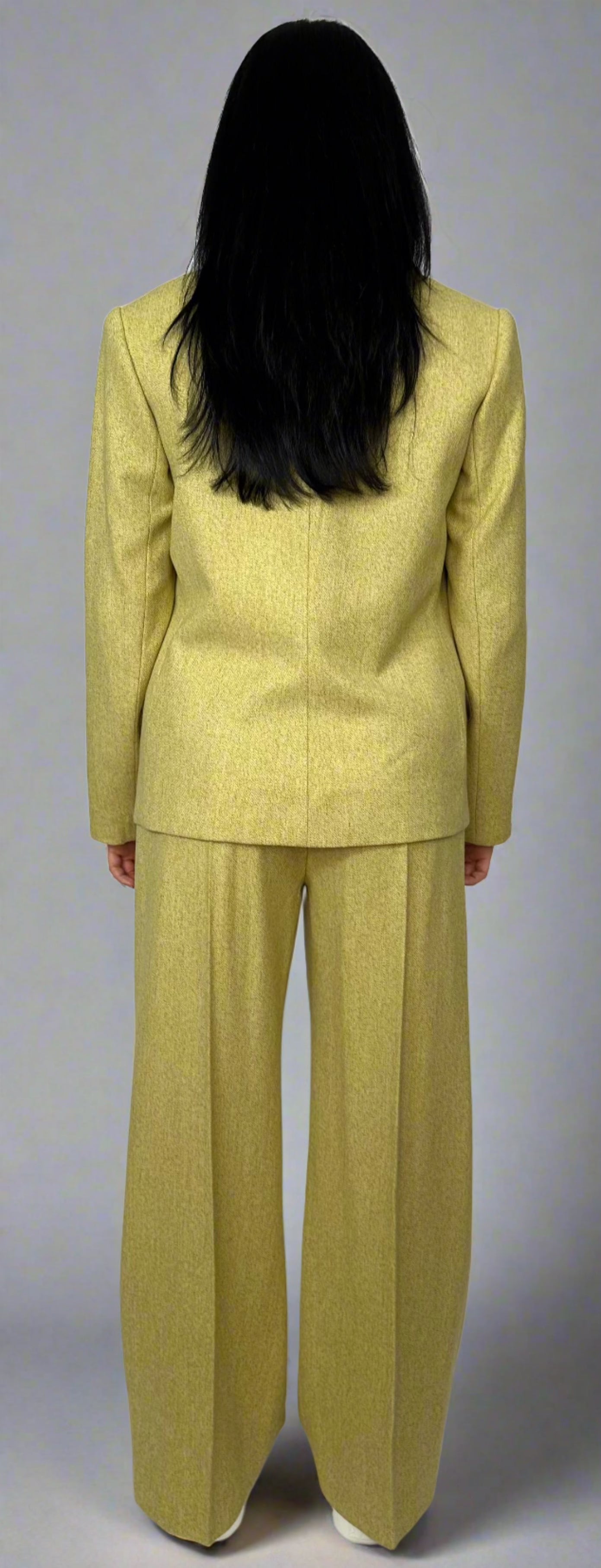 Rear view of a woman wearing a three-piece chartreuse suit by Sartorial Flair, showcasing the sleek lines of the tailored blazer, waistcoat, and wide-leg trousers, emphasizing the suit’s elegant design and fit.