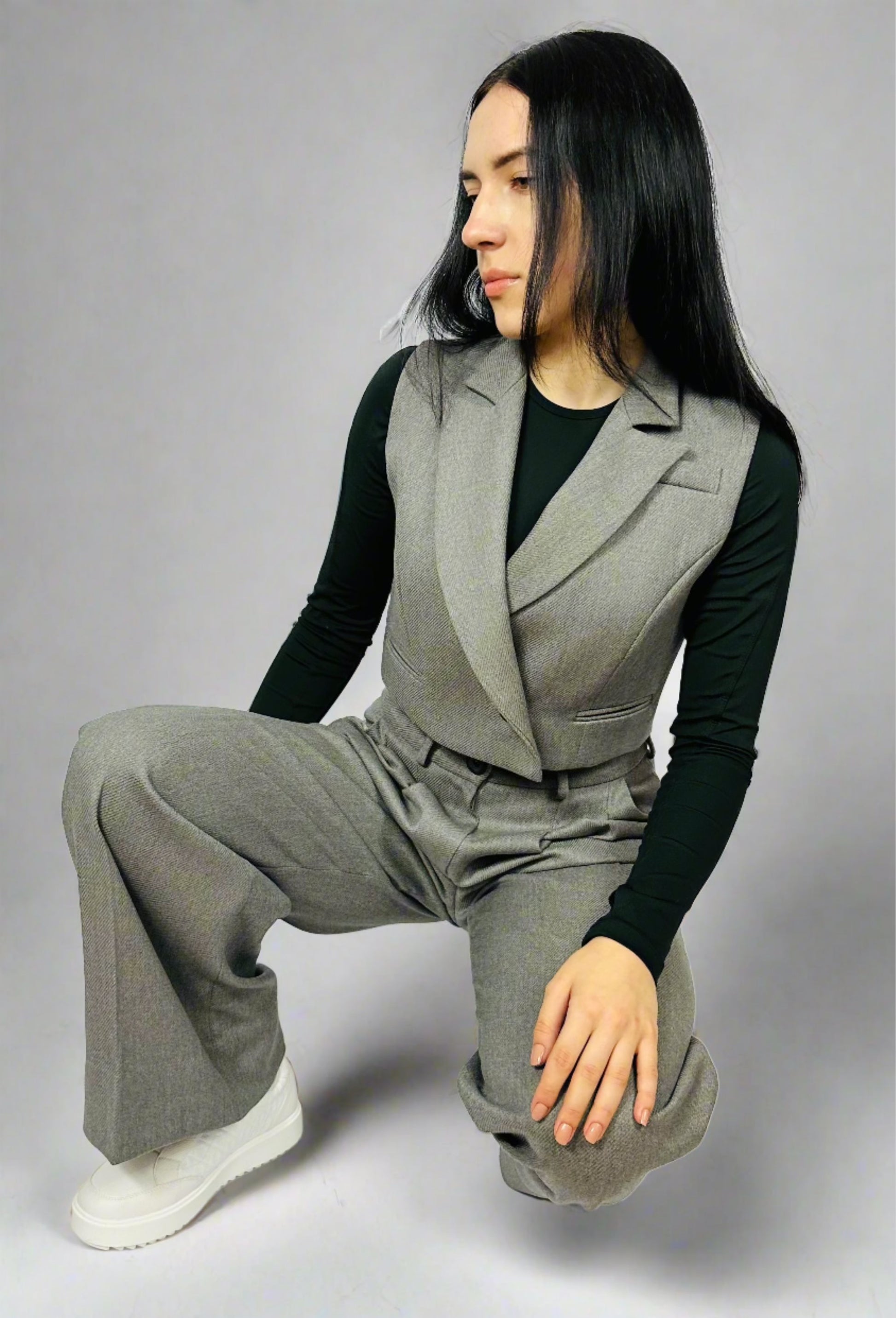 Fashionable woman squatting in a Sartorial Flair outfit, featuring a waistcoat and wide-leg trousers, exuding modern elegance against a minimalist backdrop.