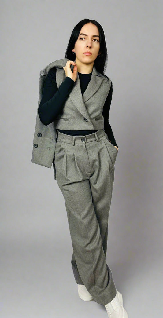 Stylish woman modeling a gray three-piece suit with a waistcoat, blazer draped over the shoulder, and wide-leg trousers, standing against a plain background.