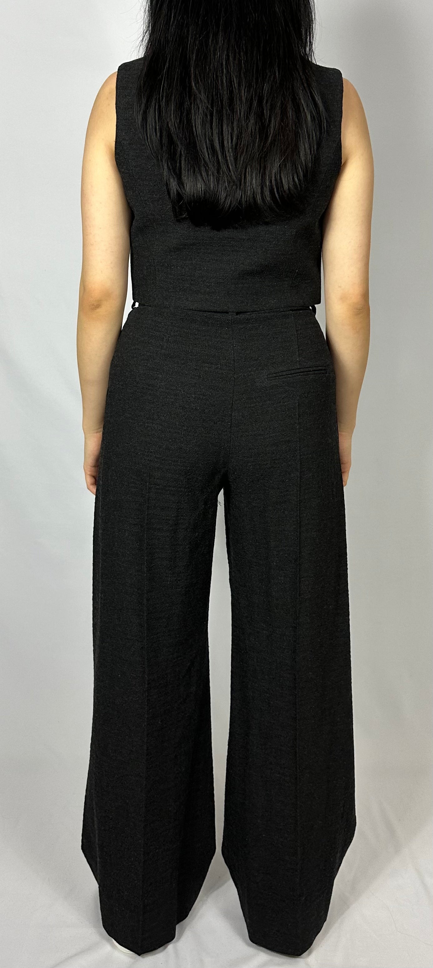 Back view of a model showcasing Sartorial Flair’s black waistcoat and matching wide-leg trousers. The waistcoat is tailored to fit closely to the body with a single-button closure. The high-waisted trousers feature a pleated front and a relaxed, wide-leg design, ensuring both style and comfort. This sophisticated ensemble by Sartorial Flair is versatile, suitable for both formal occasions and smart casual looks when paired with trainers.