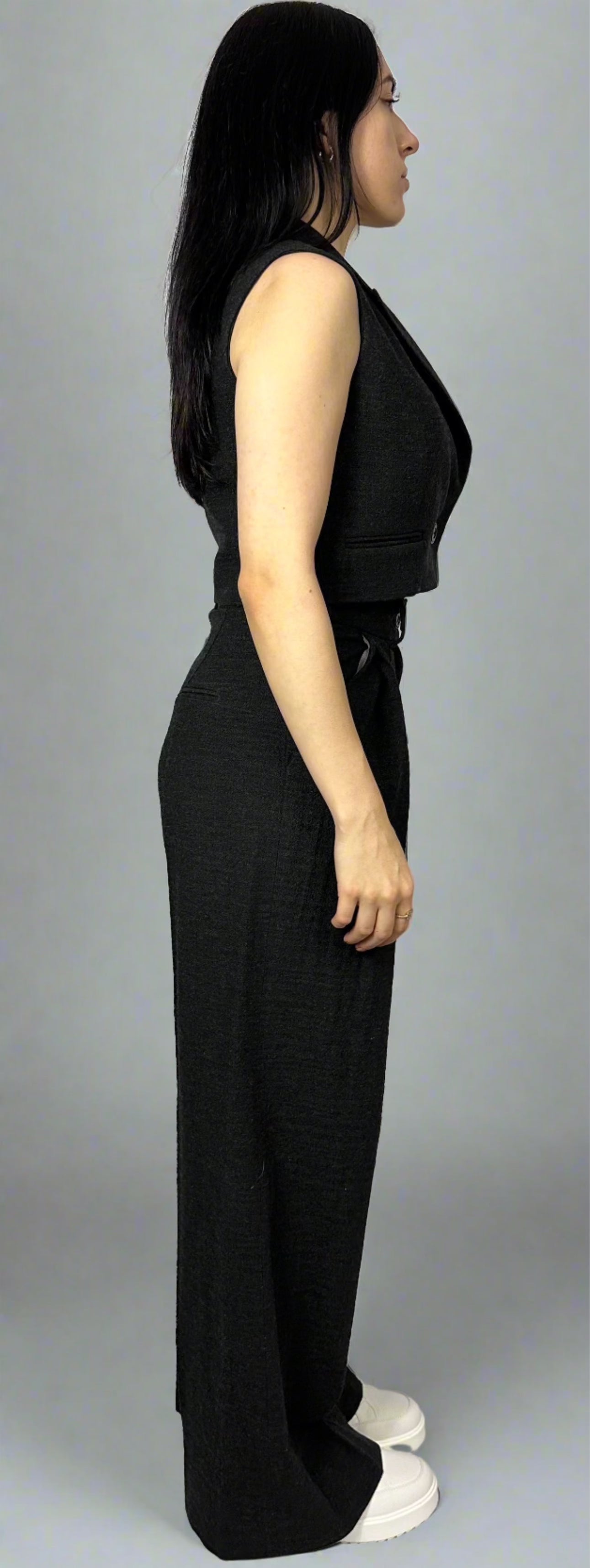 Side view of a model wearing Sartorial Flair’s black waistcoat and wide-leg trousers. The tailored waistcoat features a sleek, fitted design with a single-button closure. The high-waisted trousers have a pleated front and a relaxed fit, creating a modern and sophisticated look. Perfect for professional or formal settings, this ensemble highlights contemporary fashion elegance.