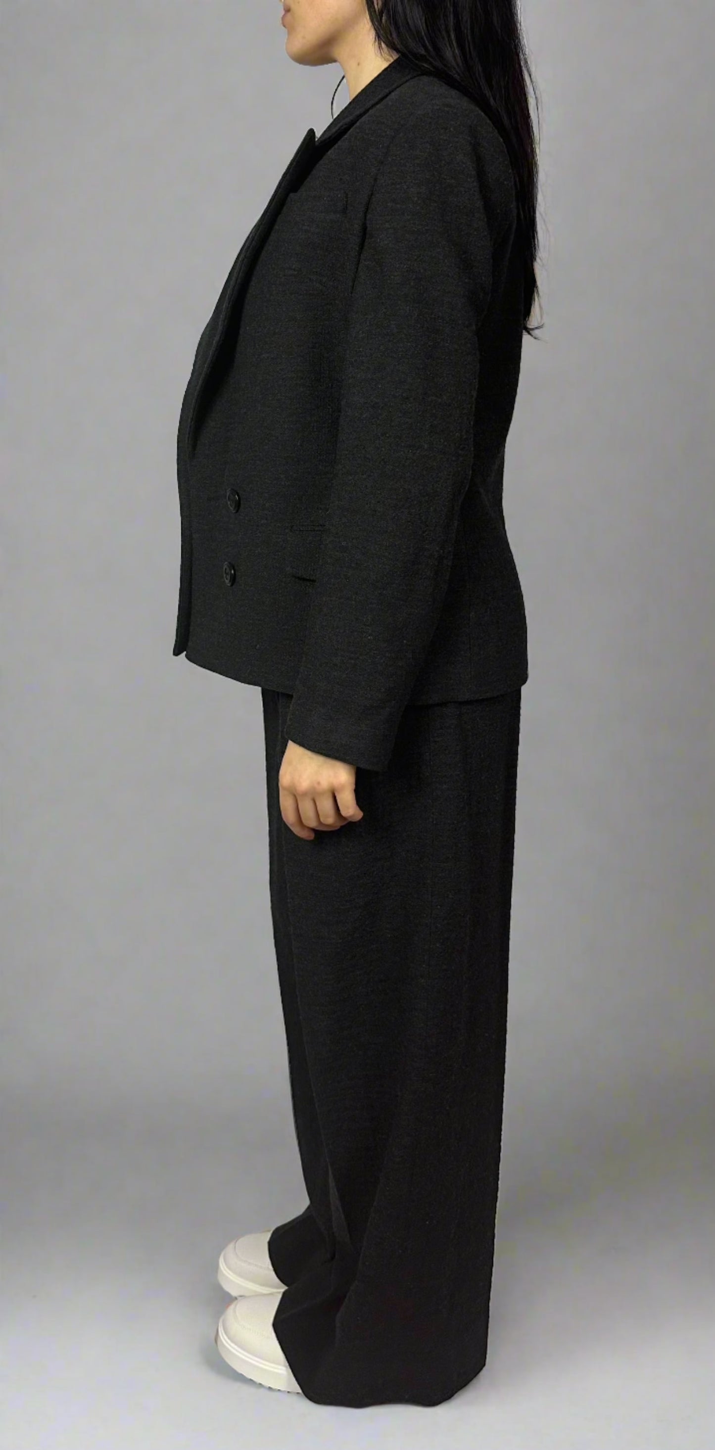 Side profile of a model showcasing Sartorial Flair’s black double-breasted blazer paired with matching wide-leg trousers. The blazer features a classic lapel design and a double-button closure, while the trousers provide a relaxed yet elegant fit. The model’s outfit embodies professional sophistication, perfect for formal business attire, representing Sartorial Flair’s commitment to contemporary yet timeless fashion.