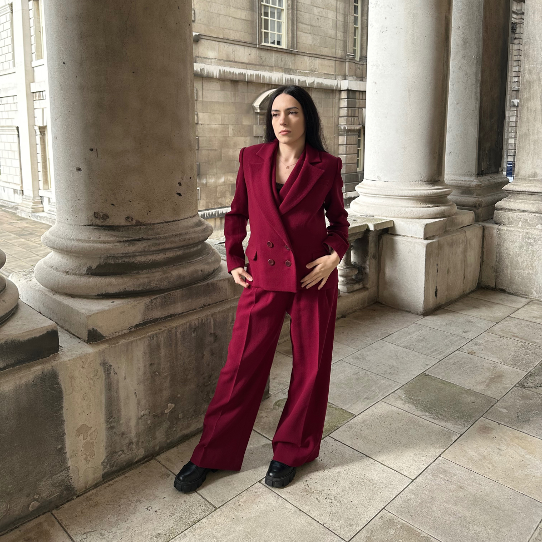 Elegant woman posing in a historic Greenwich courtyard, dressed in the Zoe deep red double-breasted suit with wide-leg trousers, from Sartorial Flair, perfect for a sophisticated and powerful fashion statement.