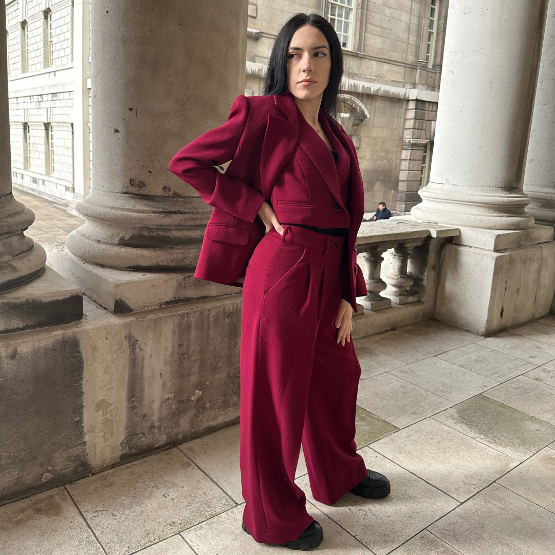 Confident woman showcasing the Zoe deep red suit, featuring a cinched waist blazer and flared trousers, posed against classical columns, embodying power and elegance from Sartorial Flair’s latest collection.